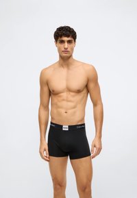 Mladý muž s krátkými tmavými vlasy, oblečený v černých boxerkách Calvin Klein, stojí proti jednoduchému světle šedému pozadí.