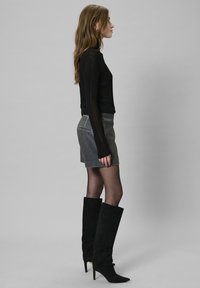 Črn rebrast turtleneck pulover, siva suknja mini iz semiša, črne škornje z visoko peto, oblečene s črnimi nogavicami; stranski pogled.