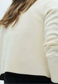 Osoba oblečená v krémovém svetru s dlouhým rukávem a černým lemem a černých kalhotách, zobrazená zezadu a z boku v úrovni pasu.