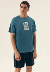 Turkusowa bawełniana koszulka z białym nadrukiem "Sunrise Surf Club", połączona z czarnymi szortami, prezentowana na neutralnym tle.