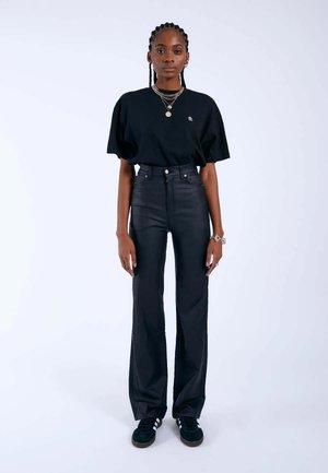 Jeune femme aux cheveux tressés, debout, portant un t-shirt noir oversize, un pantalon noir taille haute, des baskets et des colliers superposés sur fond blanc.