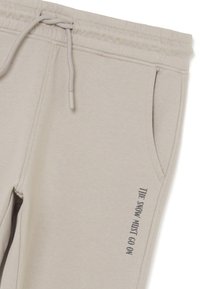 Pantalones de chándal de color beige claro con cintura elástica, cordón, bolsillo lateral y texto vertical negro que dice "LA NIEVE DEBE CONTINUAR."