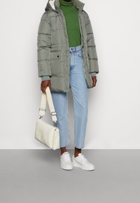 Abrigo acolchado verde con capucha, bolsillos delanteros y puños elásticos, combinado con jeans azul claro, zapatillas blancas y un bolso crema.