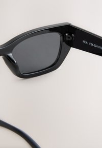 Schwarze Sonnenbrille mit glänzendem Kunststoffrahmen, rechteckigen Gläsern und einem dezenten Designdetail am Bügel. Dahinter ein dünnes schwarzes Band.
