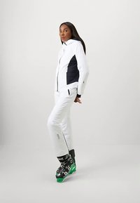 Γυναίκα που φοράει ένα λευκό μπουφάν και παντελόνι σκι με μαύρες και πράσινες μπότες σκι, στέκεται σε ένα ουδέτερο στούντιο.