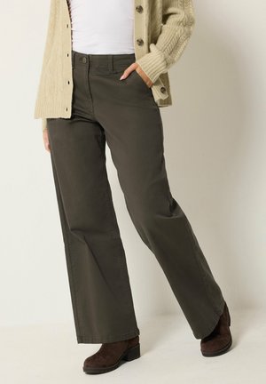 Donna che indossa pantaloni a gamba larga color oliva scuro, cardigan beige lavorato a maglia, top bianco e stivaletti marrone scuro, in piedi con una mano in tasca.