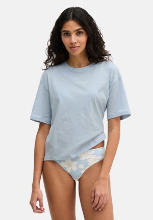 Vrouw met lang krullend haar, gekleed in een lichtblauw oversized T-shirt en lichtblauwe bikinibroek met bloemenprint, tegen een effen achtergrond.