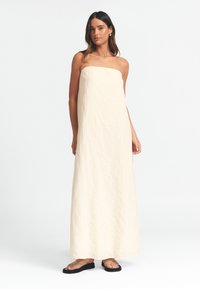 Maksi obleka brez naramnic v svetlo bež teksturiranem materialu, ki ima vertikalne rebraste vzorce. V kombinaciji s preprostimi črnimi sandalami.