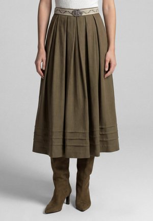 Vrouw draagt een olijfgroene geplooide midi-rok met decoratieve riem en bijpassende suède laarzen met hak tegen een effen achtergrond.