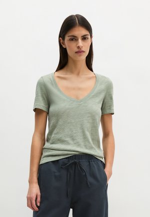 Vrouw met lang donker haar die een lichtgroen T-shirt met V-hals en korte mouwen draagt en een donkere broek met trekkoord, staand met één hand in de zak.