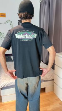 Oseba, oblečena v črno majico Timberland in kapo, stoji z rokami na bokih v spalnici, v ozadju postelja in zavese.