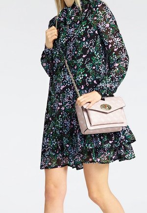 Frau in einem schwarzen Blumenkleid, die eine hellrosa Umhängetasche mit einer Kettenriemen und einem goldenen Verschluss hält.