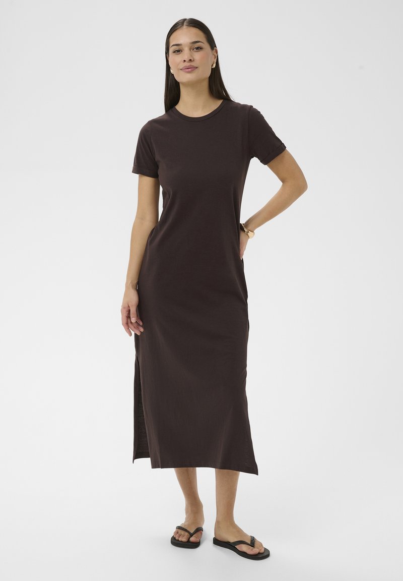 Femme aux cheveux longs portant une robe midi marron foncé à manches courtes avec une fente latérale et des tongs noires, debout devant un fond uni.