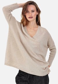 Pull beige ample à col en V, fabriqué en tissu tricoté doux avec poignets et ourlet côtelés. La texture semble lisse avec des variations subtiles.