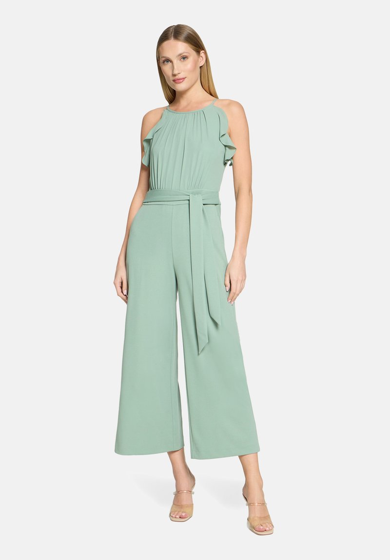 Kvinde iført ærmeløs lysegrøn heldragt med flæsedetaljer og taljebånd, stående foran en neutral studiebaggrund.