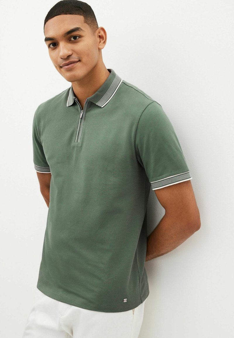 Grøn polo-shirt med tekstureret stof, korteærmer, lynlås krave og stribede detaljer på kraven og ærmerne. Lille logo detalje i sømmen.