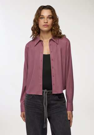 Camicia a maniche lunghe accorciata in malva, realizzata in un tessuto liscio, con chiusura a bottoni e colletto classico, abbinata a jeans scuri.