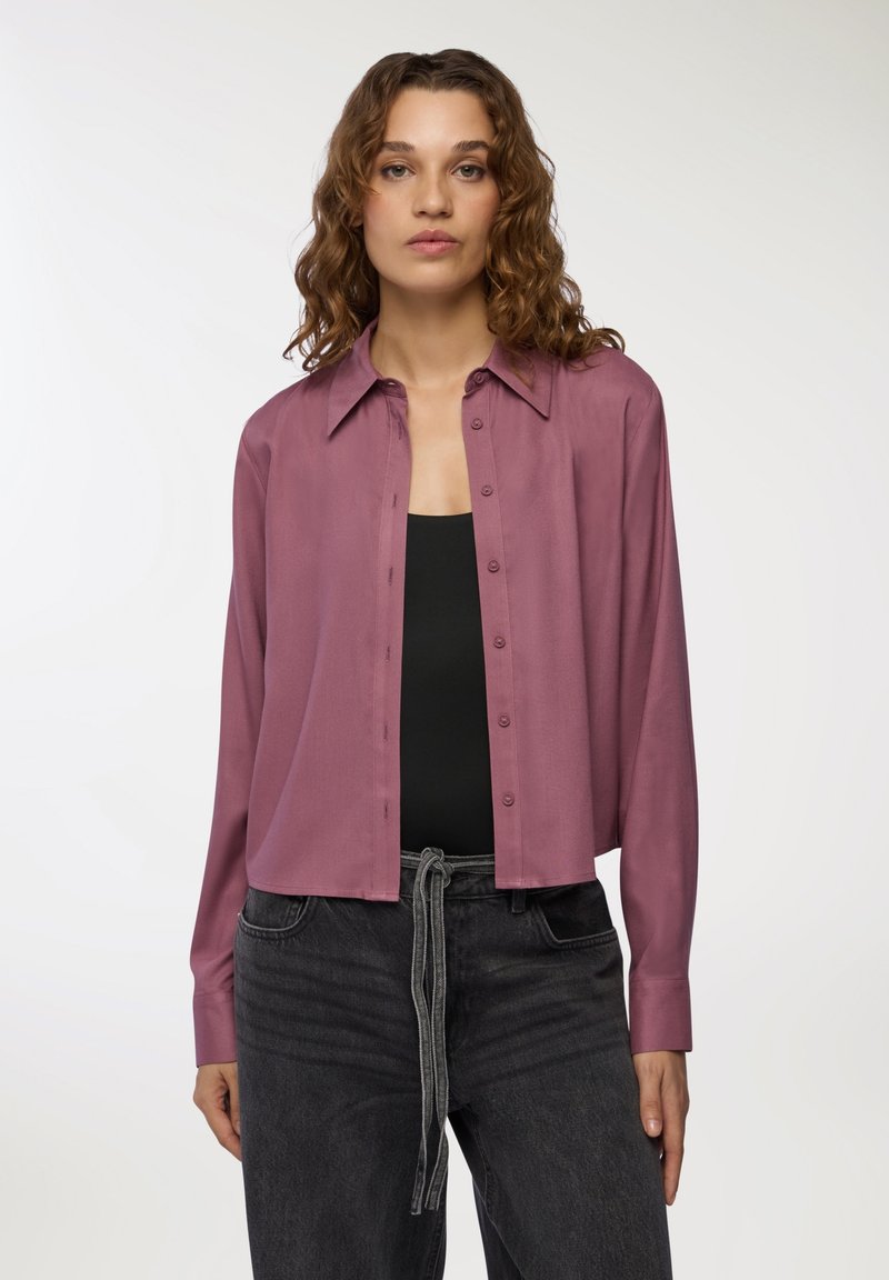 Camicia a maniche lunghe accorciata in malva, realizzata in un tessuto liscio, con chiusura a bottoni e colletto classico, abbinata a jeans scuri.