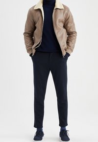 Veste en similicuir marron avec un col en mouton crème, associée à un pull et un pantalon bleu marine. Le modèle porte des baskets bleu marine ; look décontracté.