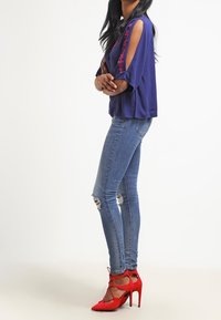 Paarse blouse met uitgesneden schouders en bloemenborduurwerk, gecombineerd met distressed lichtblauwe jeans en felrode hoge hakken.