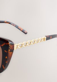 Lunettes de soleil écaille de tortue avec un détail chaîne en or sur la branche du côté, sur un fond neutre.