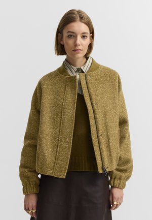 Frau mit kurzem blonden Haar, die eine grünlich-braune strukturierte Jacke über einem dunklen Pullover und einem gestreiften Hemd mit Kragen trägt, steht vor einem einfachen Hintergrund.