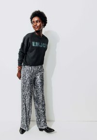 Sweat-shirt noir avec "ENJOY" en lettres réfléchissantes, associé à un pantalon large en argent scintillant. Des bottes à talons aiguilles noires viennent compléter la tenue.