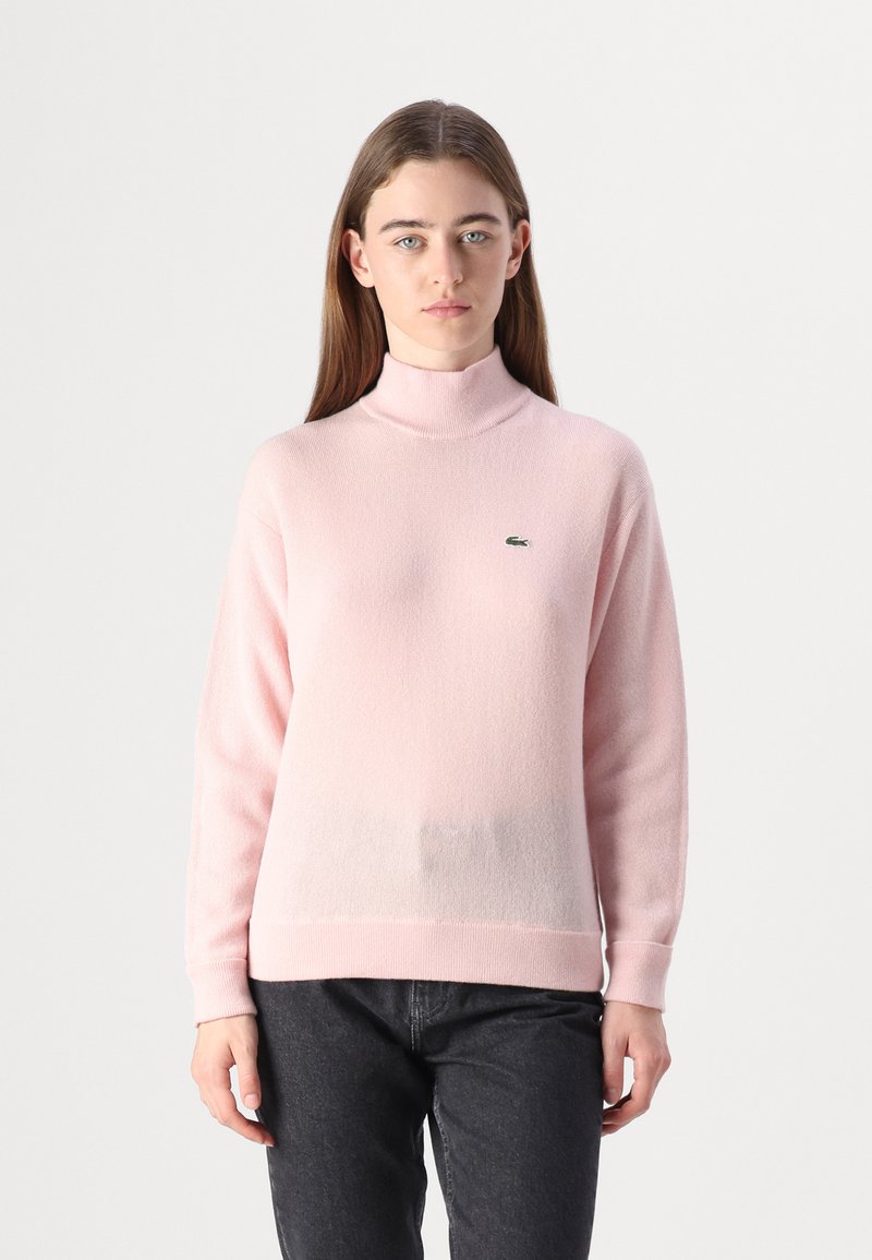 Ljusrosa mock turtleneck-tröja gjord av mjukt tyg, med en liten grön logotyp på bröstet samt ribbade muddar och fåll.