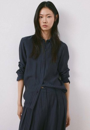 Joven con cabello largo y oscuro, vistiendo una camisa azul marino de rayas y pantalones a juego, de pie contra una pared lisa y clara.