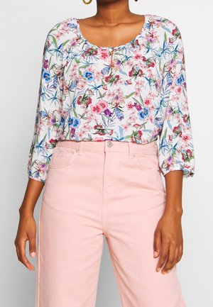 Vrouw die een witte blouse met roze, blauwe en paarse bloemen draagt, gecombineerd met hooggetailleerde lichtroze broek en gouden oorbellen in de vorm van ringen.