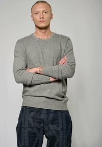 Pull gris à manches longues avec des poignets et un ourlet en côtes, associé à un pantalon foncé à motifs avec des formes géométriques et des points clairs.