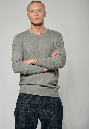 Pull gris à manches longues avec des poignets et un ourlet en côtes, associé à un pantalon foncé à motifs avec des formes géométriques et des points clairs.