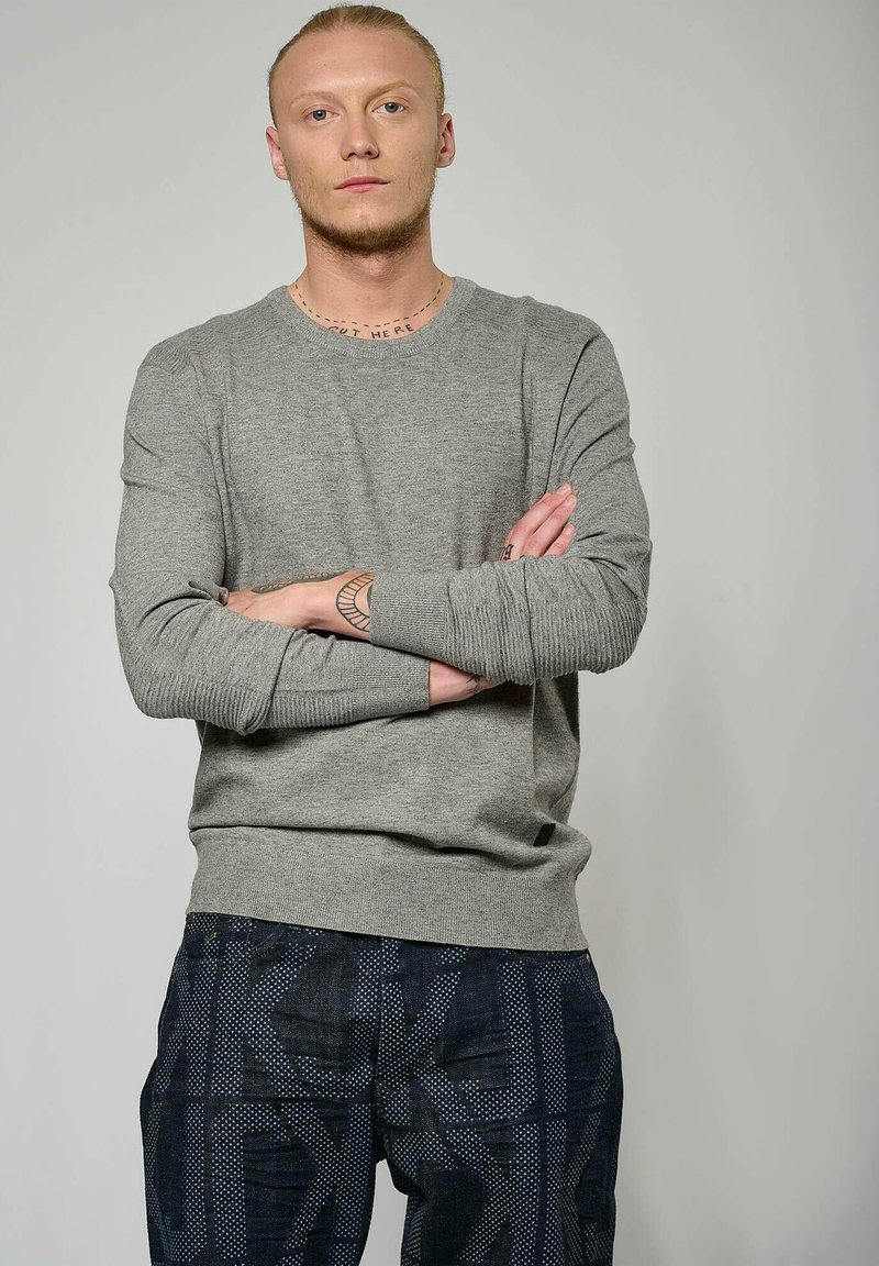 Pull gris à manches longues avec des poignets et un ourlet en côtes, associé à un pantalon foncé à motifs avec des formes géométriques et des points clairs.