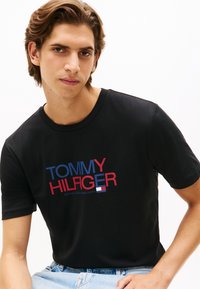 Μαύρο βαμβακερό T-shirt με στάμπα "TOMMY HILFIGER" σε κόκκινο και μπλε, στρογγυλή λαιμόκοψη, κοντά μανίκια και άνετη εφαρμογή.