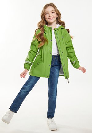 Giacca impermeabile verde con cappuccio, bottoni a pressione e tasche laterali. Indossata con jeans blu e sneakers bianche; superficie liscia e finitura lucida.