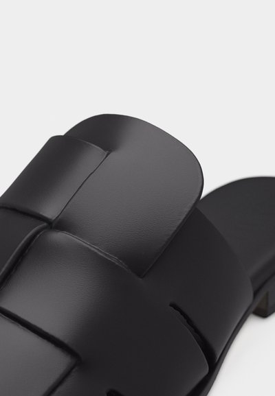 Μαύρο δερμάτινο σανδάλι τύπου slip-on με φαρδιές διασταυρούμενες λουριά και χαμηλό τακούνι block, σε λευκό φόντο.