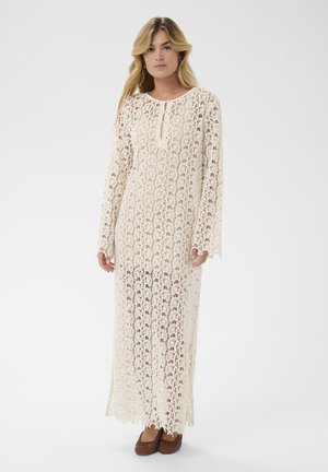 Lang, cremefarvet hækle kjole med rund udskæring, lange ærmer og åbent mønster. Kendt for sin delikate blonde tekstur og flydende design.