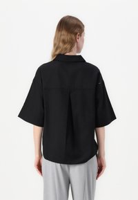 Camisa preta de manga curta com colarinho, apresentando um plissado nas costas e um ajuste descontraído. Feita de tecido suave com detalhes de design mínimos.