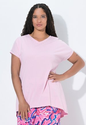 Femme aux longs cheveux tressés porte un haut rose clair à manches courtes et un pantalon aux motifs vifs rose, bleu et blanc, debout avec la main sur la hanche.