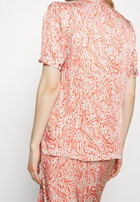 Košile s krátkým rukávem z lehké látky s abstraktním vzorem v korálové a bílé barvě; detail představují ohrnuté rukávy a kulatý lem.