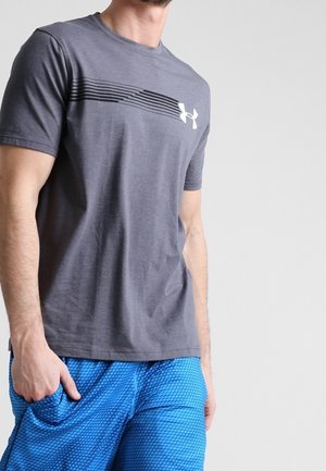 Mężczyzna w szarej koszulce Under Armour z logo oraz niebieskich sportowych spodenkach z teksturą, stojący na tle jednolitego jasnego tła.