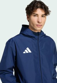 Ναυτικό μπουφάν με κουκούλα, κατασκευασμένο από λείο ύφασμα, διαθέτει μπροστινό φερμουάρ και άσπρο λογότυπο Adidas στο αριστερό στήθος. Χωρίς σχέδια ή επιπλέον λεπτομέρειες.