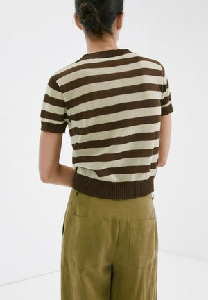 Persoană cu părul prins în coc, purtând un pulover cu mâneci scurte în dungi maro și bej și pantaloni verde măsliniu, văzută din spate.