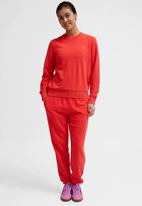 Czerwona bluza i pasujące spodnie dresowe wykonane z miękkiego materiału, z okrągłym dekoltem. Z przodu na piersi znajduje się małe logo, a po bokach kieszenie.