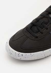 Zapatilla deportiva negra con parte superior texturizada, puntera redondeada y suela moteada en blanco. Los cordones y las costuras son negros, creando un diseño unificado.