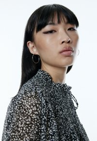 Zwart-wit gestreepte blouse met een gerimpelde halslijn, geplooide textuur en mouwen. Model draagt ronde zilveren ringoorbellen.