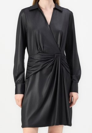 Kobieta ubrana w czarną satynową sukienkę z długim rękawem, kołnierzykiem i marszczonym detalem w talii, stojąca na jednolitym białym tle.