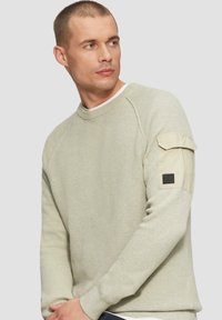 Jasnozielony sweter o fakturze z okrągłym dekoltem, z podwijanym wykończeniem i naszywaną kieszenią na lewym rękawie.