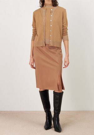 Vrouw draagt een camel-kleurige cardigan en bijpassende top, met een camel-kleurige rok tot op de knie met zijsplit en zwarte kniehooge laarzen.