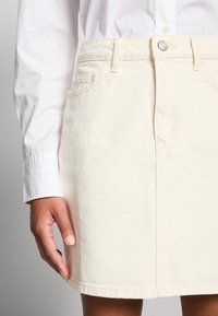 Personne portant une chemise blanche à manches longues et une jupe en denim crème avec des poches avant visibles et une fermeture à boutons.